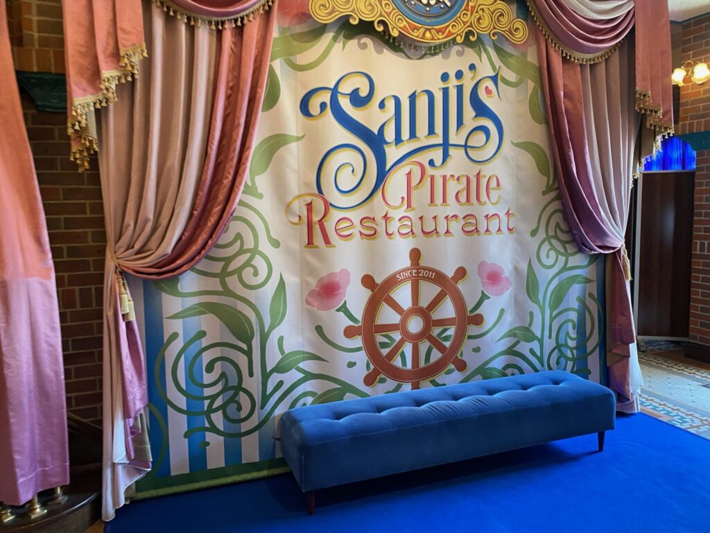 「Sanji's Pirate Restaurant SINCE 2011」のロゴと舵輪が描かれた撮影用の背景幕。前に青いベンチと青いカーペット