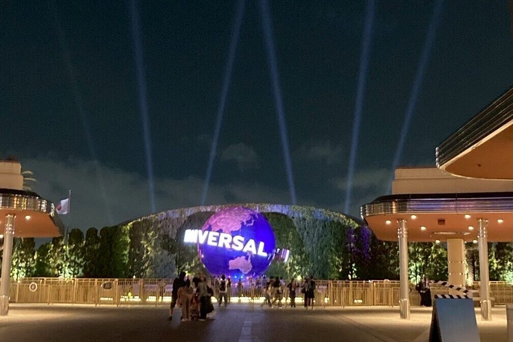 夜空にサーチライトの光が伸びる中、紫色にライトアップされたユニバーサル・グローブと、その前を歩く人たち