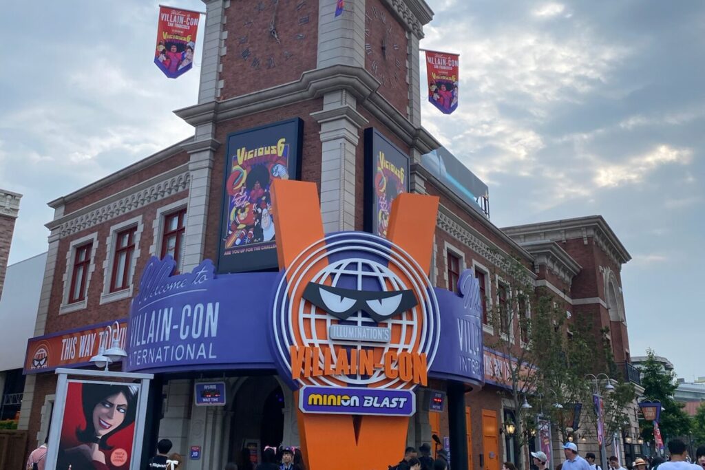 レンガの建物の前に「VILLAIN-CON」の看板とオレンジ色のV字のロゴが立つ、ミニオン・ハチャメチャ・ミッションの入口